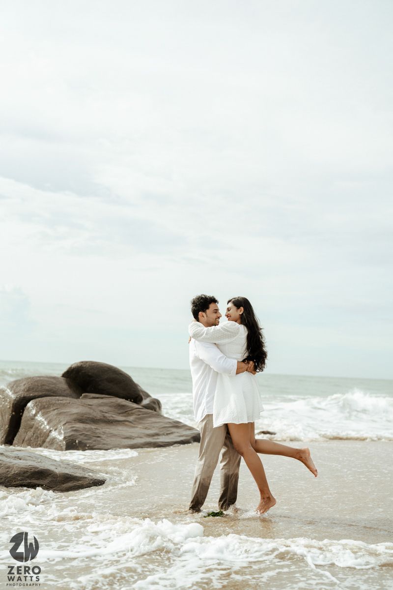 A couple in light colored clothing embraces on a rocky beach with waves crashing nearby.