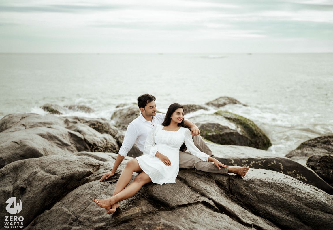Couple in white sits together on large coastal rocks overlooking the ocean.