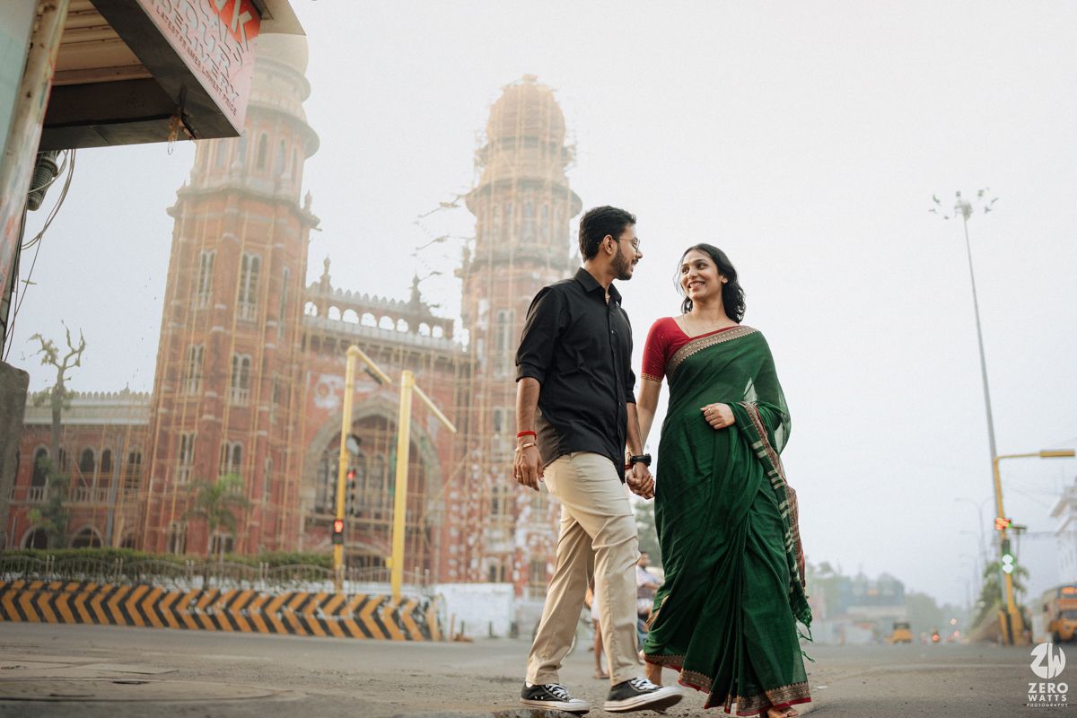 A couple in casual and traditional attire poses in front of a grand historical building with twin towers.