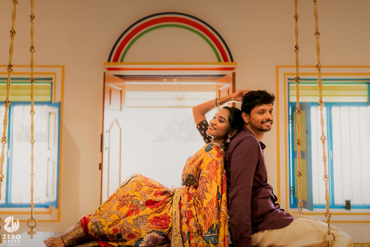 Couple in traditional Indian clothing pose back-to-back against colorful window with rainbow arch.
