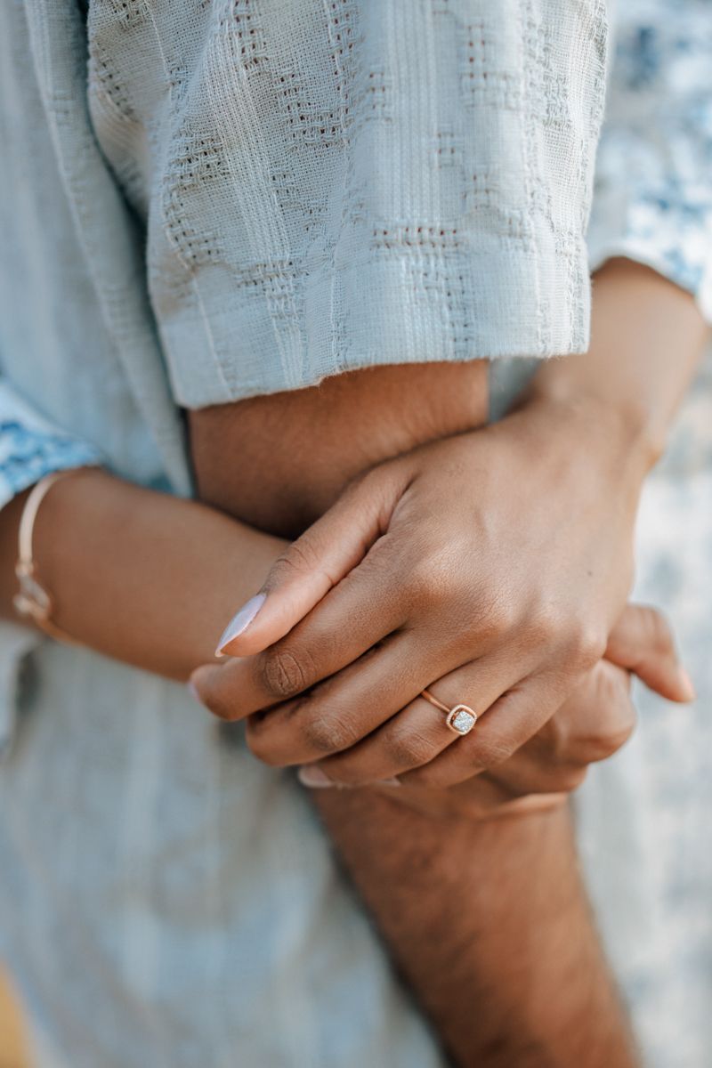 Close up of hands wearing an engagement ring and delicate bracelet against light blue textured fabric.