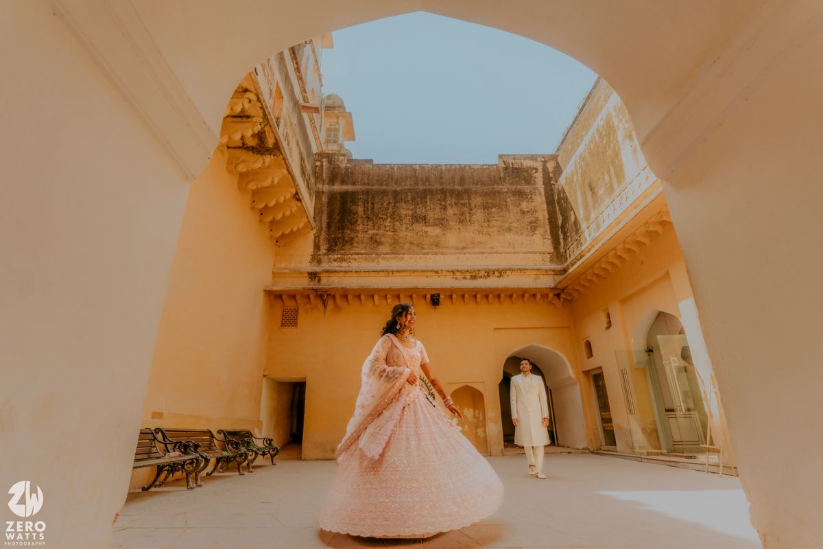 A person wearing a flowing pink gown poses in a traditional Indian palace courtyard with intricate architectural details.