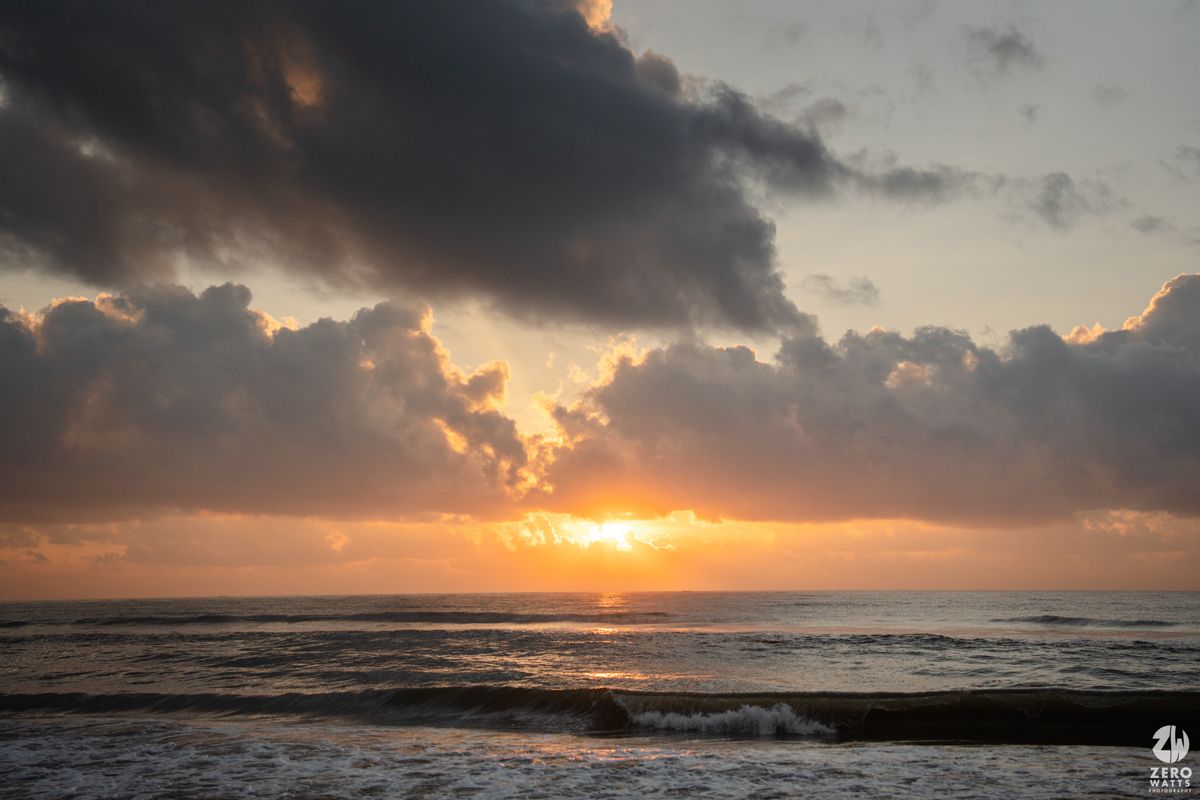 Dramatic ocean sunset with golden sun peeking through dark clouds over rolling waves.