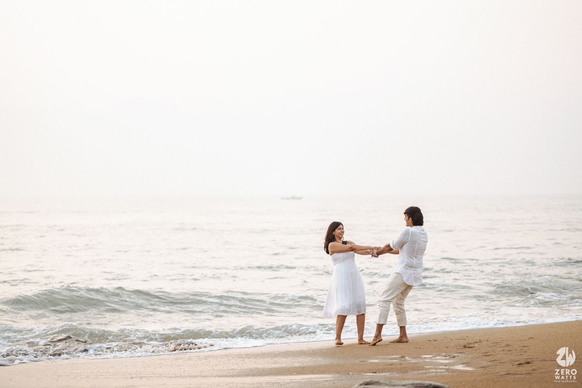 A couple in white outfits dance together on a serene beach at sunset with ocean waves rolling in the background.