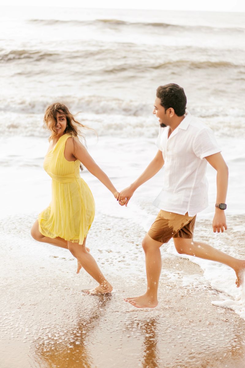 Couple runs playfully together on beach shoreline with waves crashing, woman in yellow dress and man in white shirt.