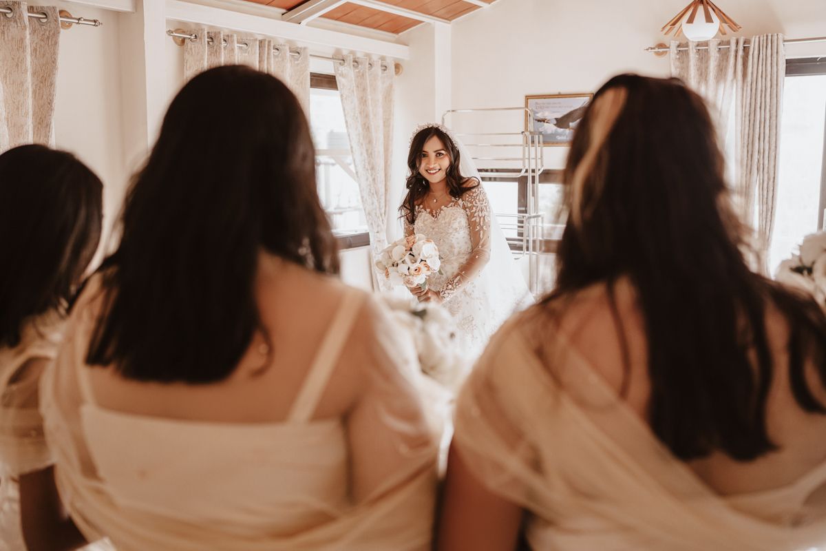 A bride in a white wedding gown sits in a sunny room with her bridal party wearing matching robes.