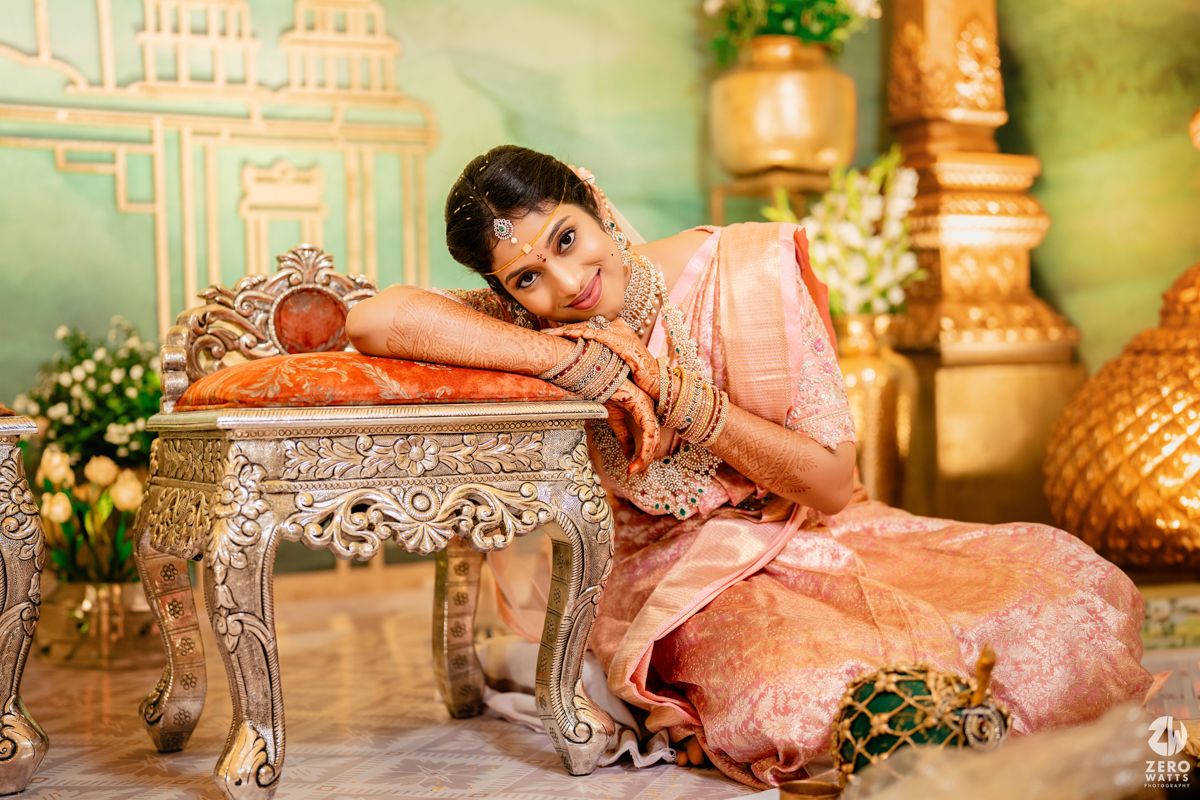 A person in a peach-colored traditional dress poses elegantly beside an ornate silver table in a decorated setting.