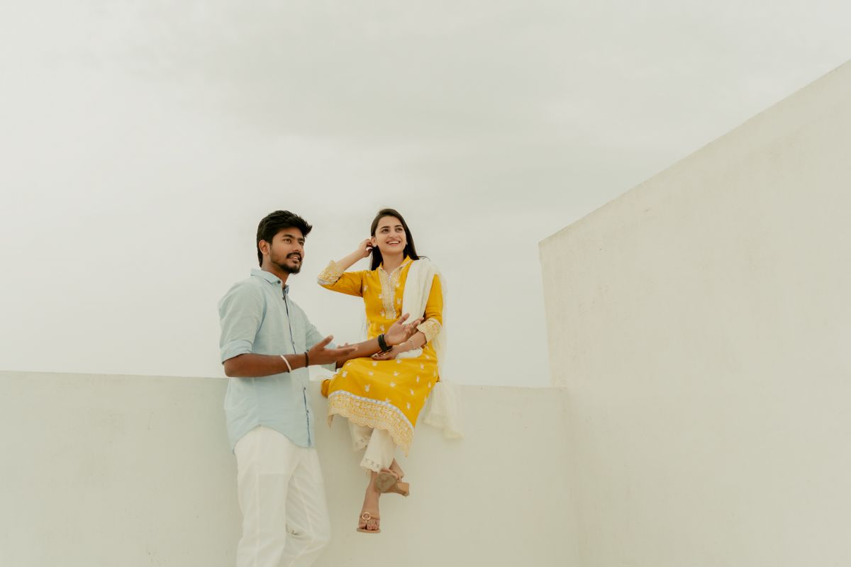 A couple standing in a minimalist white space with one wearing a yellow dress and the other in light blue attire.