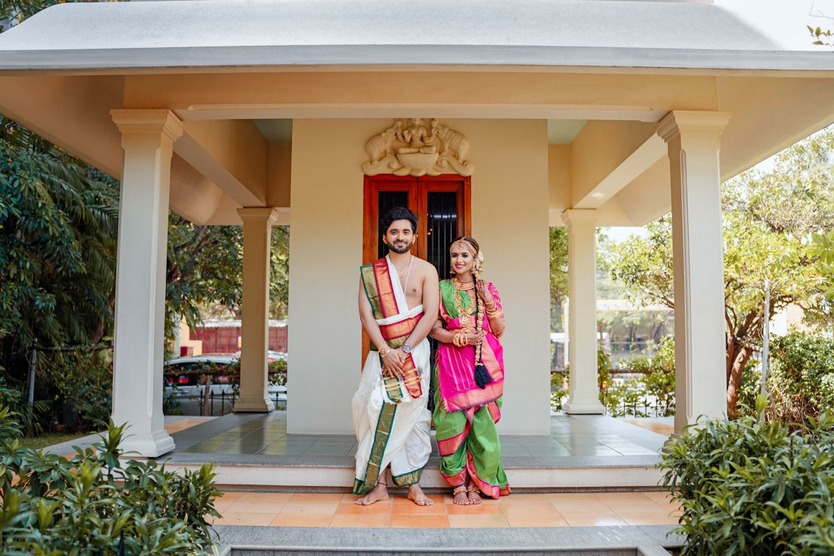 A couple in traditional Indian attire poses on the porch of a cream-colored building with white pillars.