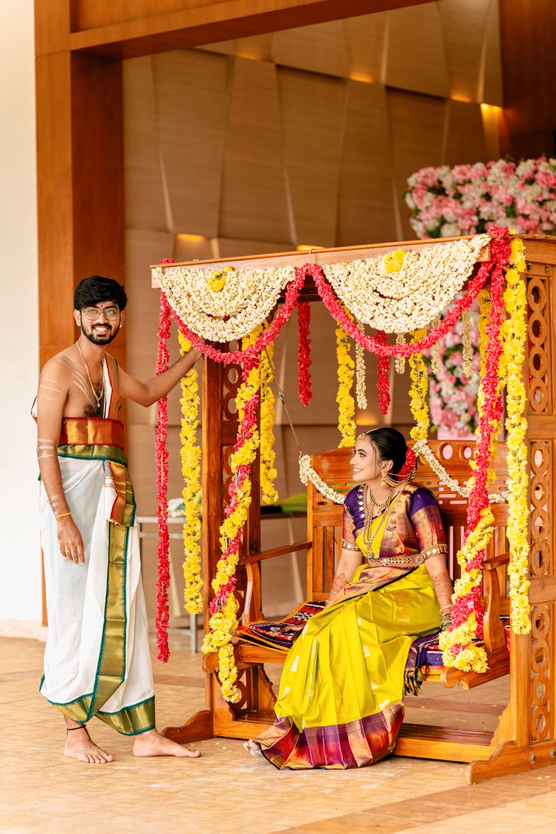 Traditional South Indian couple in wedding attire pose with decorative swing adorned with marigolds and floral arrangements.