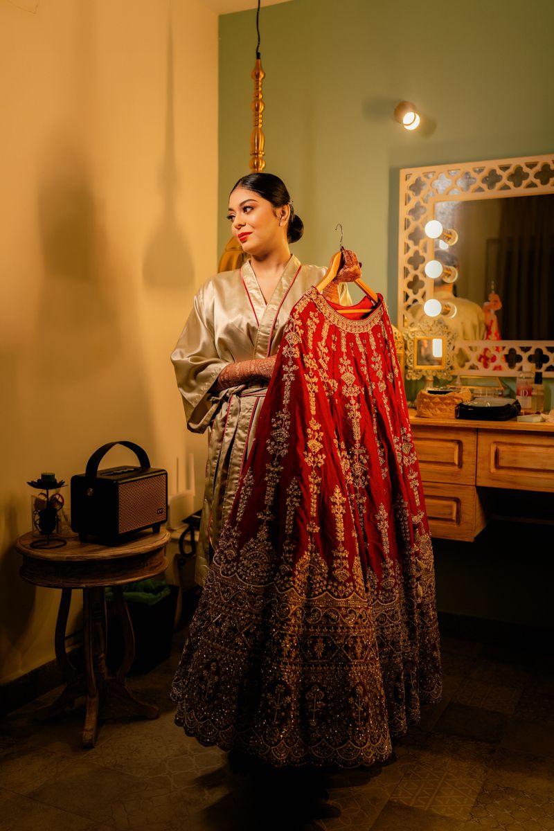 A red and gold embroidered bridal lehenga hangs on display next to a dressing table with vanity lights in warm lighting.