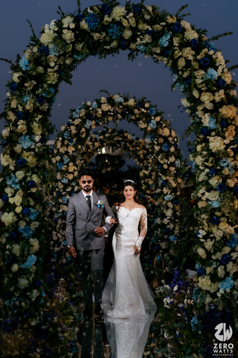 Elegant wedding couple stands beneath beautiful floral arches decorated with white roses and blue flowers at dusk.