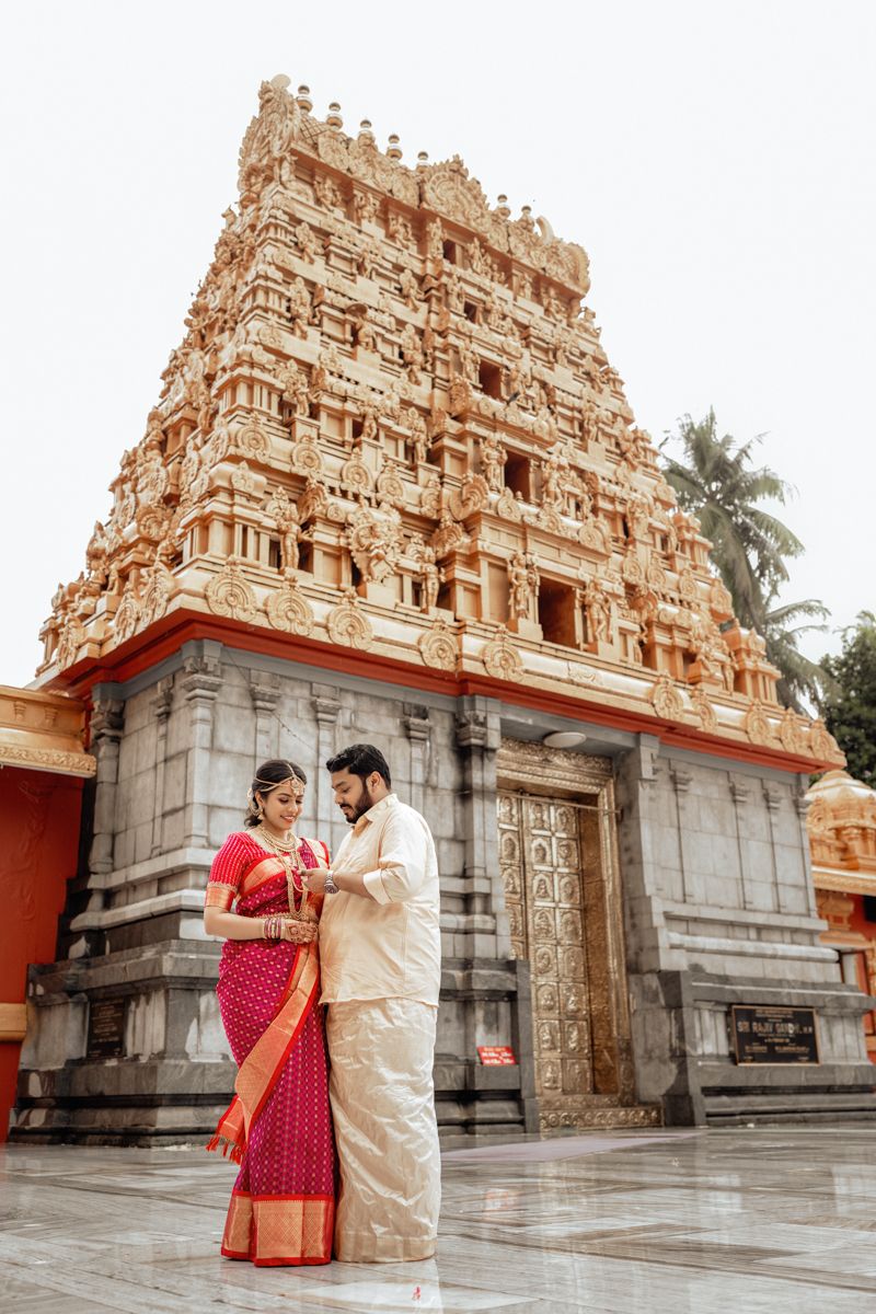 A couple in traditional South Indian attire pose in front of a majestic temple gopuram with intricate architectural details.