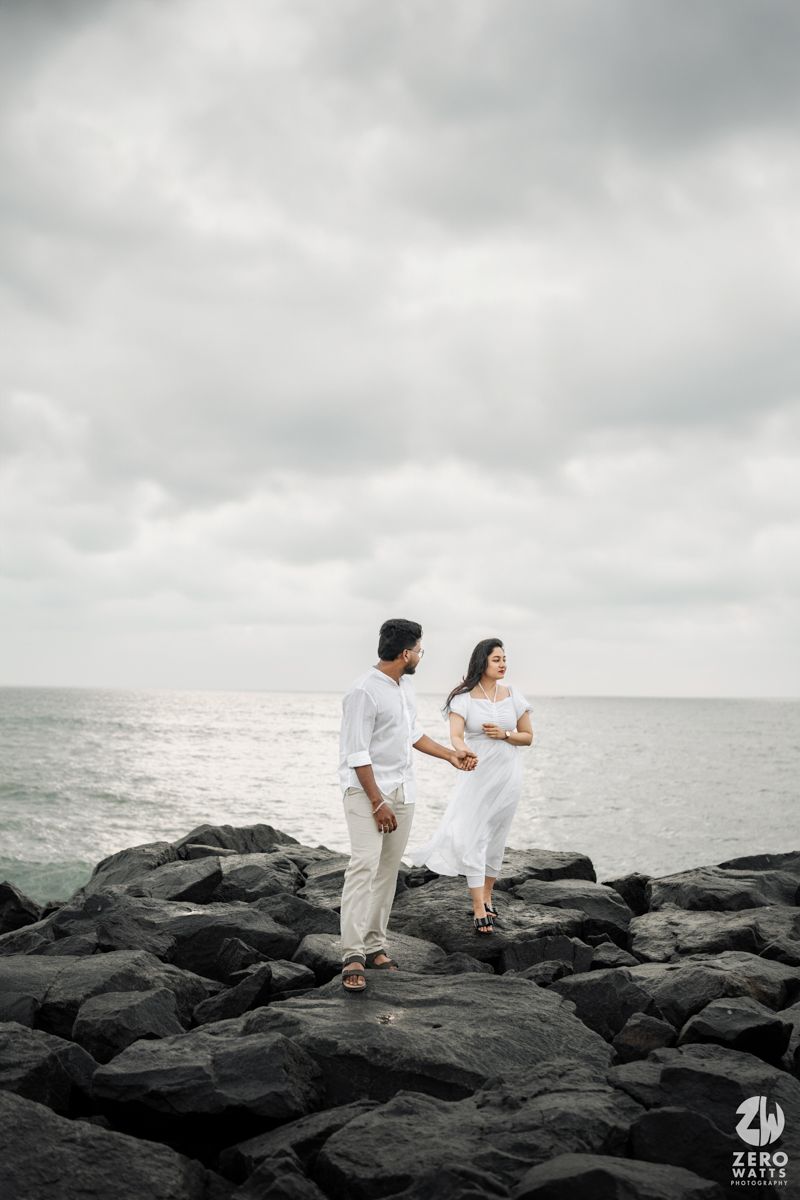 Couple in white clothing walking on black coastal rocks against moody ocean backdrop.
