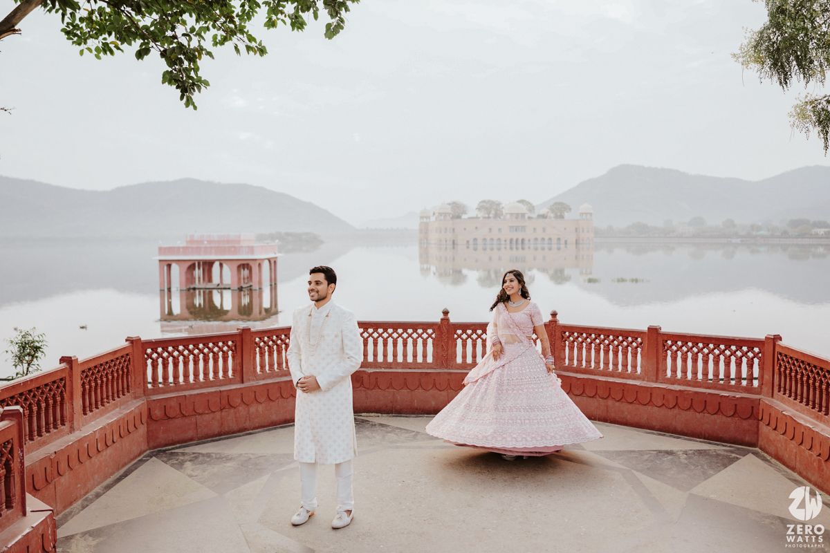 A couple in white attire stands on a red-railed terrace overlooking a misty lake and palace in the background.