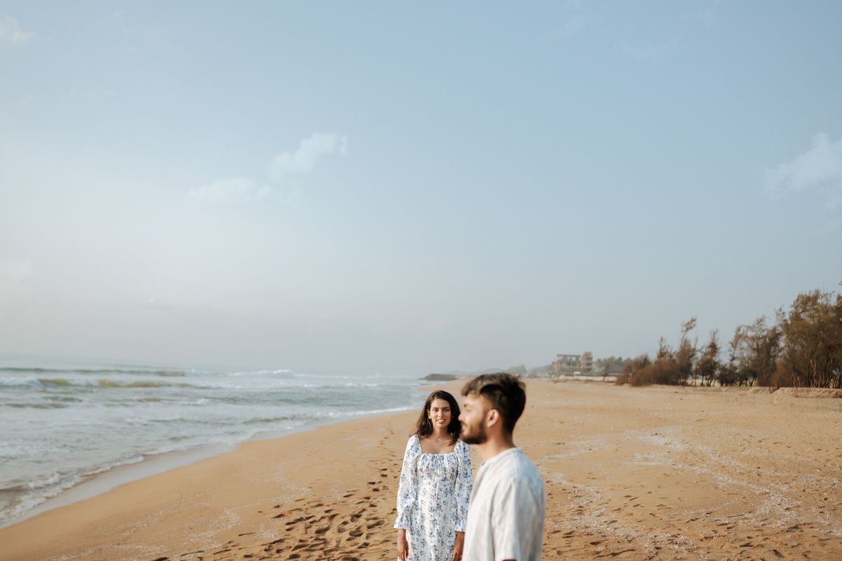 A couple in casual attire standing together on a sandy beach with ocean waves and trees visible in the background.
