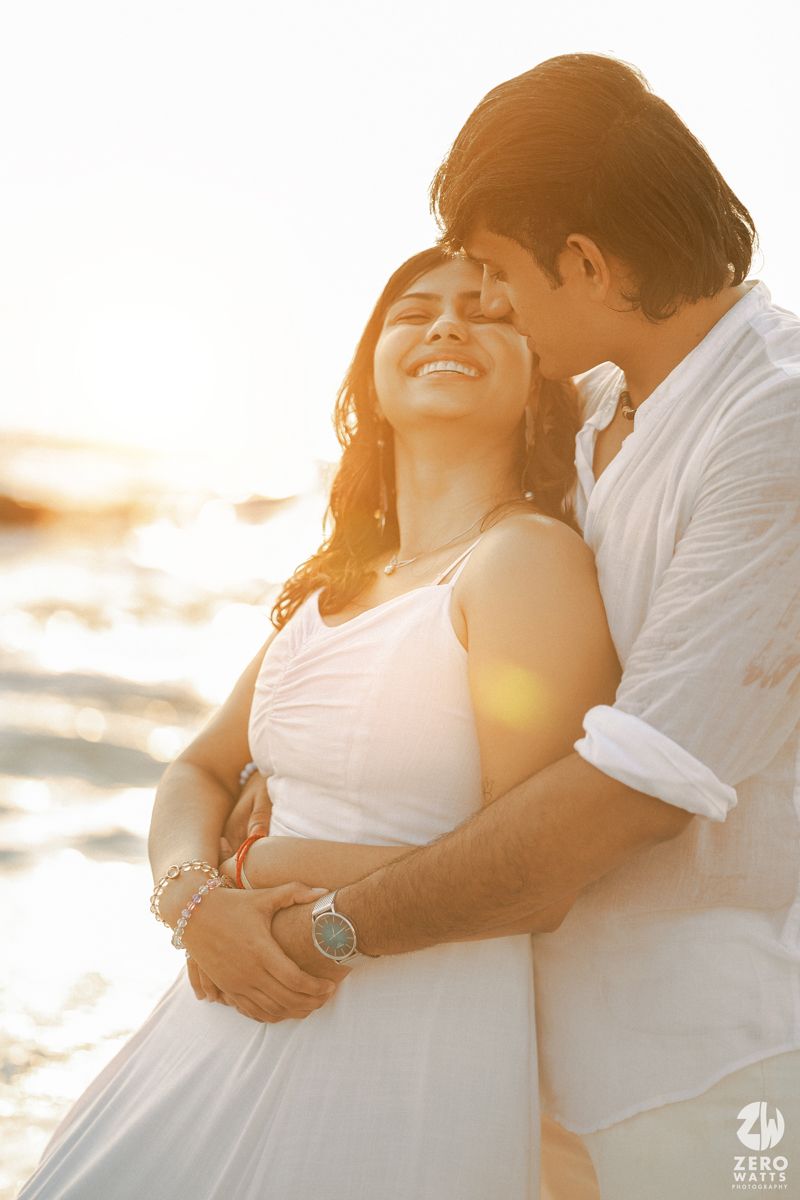 Romantic couple embraces at sunset on a beach in white outfits with golden sunlight streaming through.