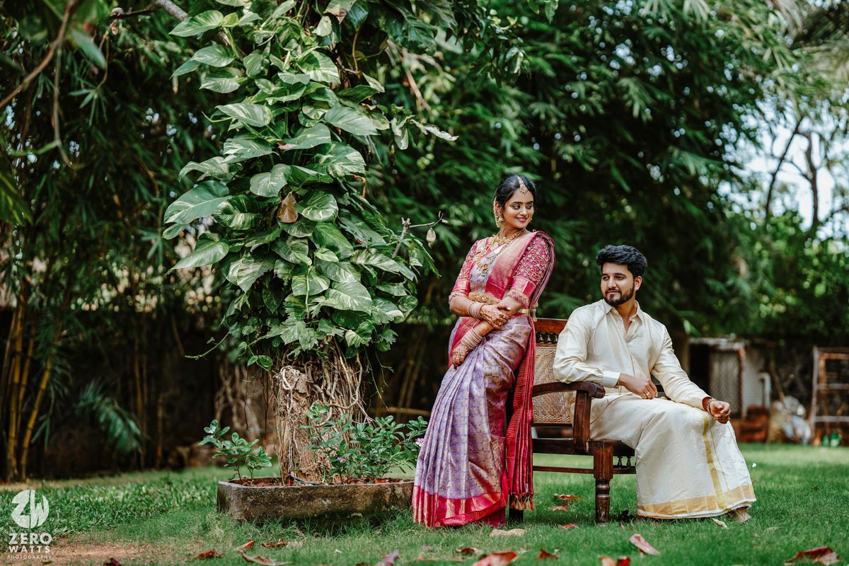 A couple in traditional Indian attire pose in a lush garden setting with green foliage in the background.