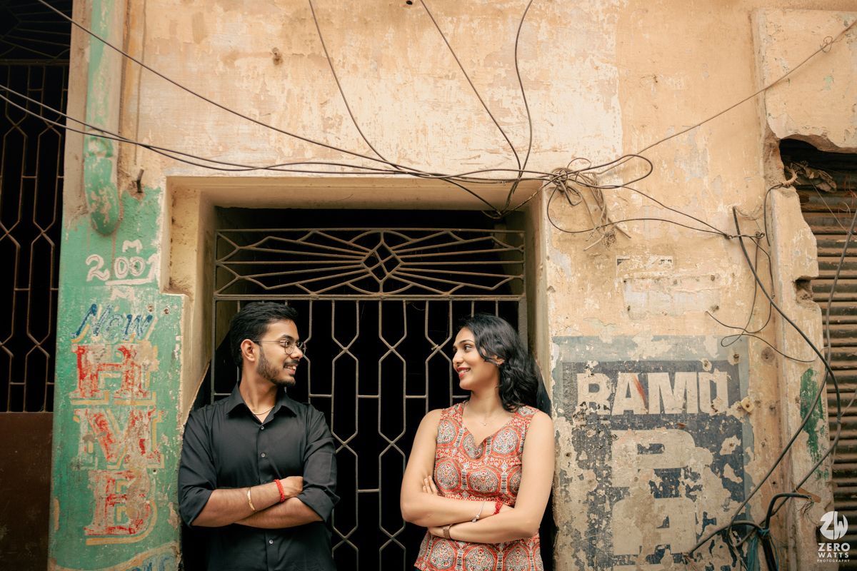A couple stands before a weathered wall with graffiti and iron grates in an urban street scene.