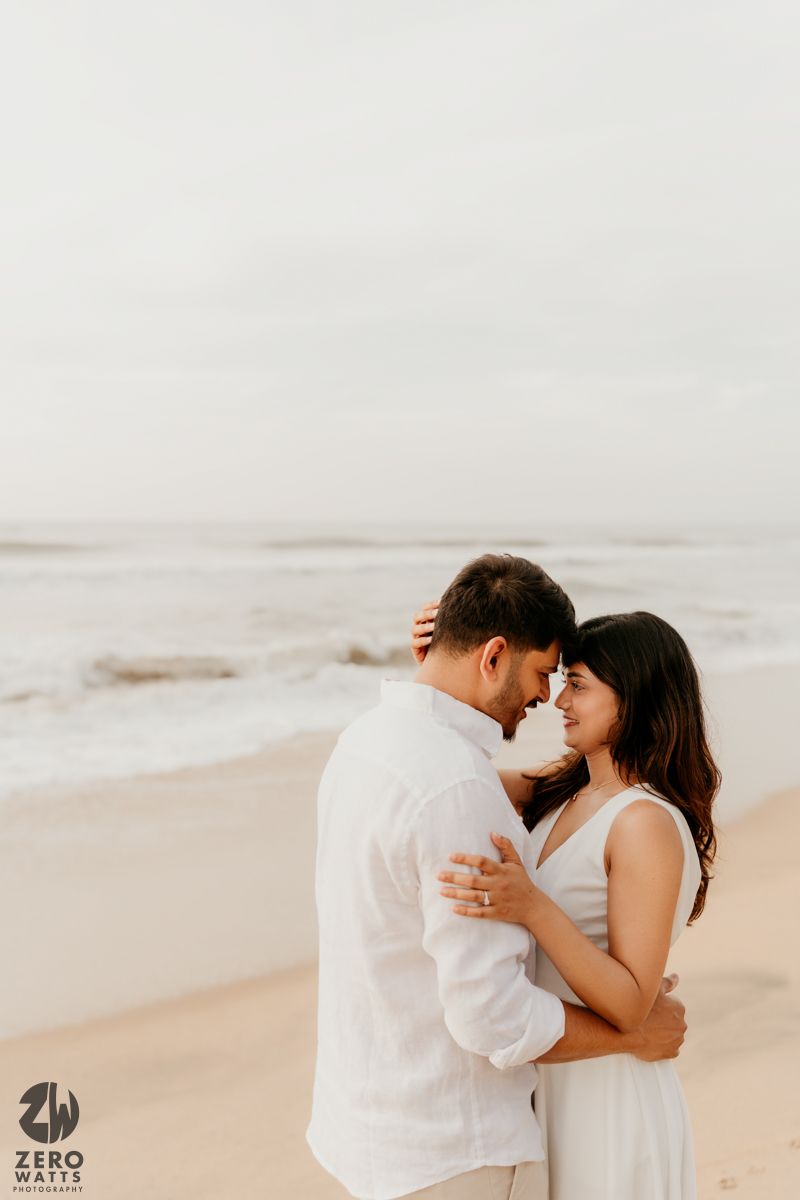 A romantic couple embraces on a misty beach at sunset wearing white outfits.
