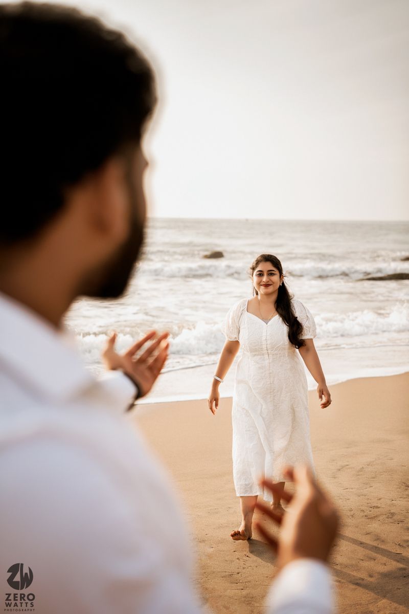 Candid beach photography showing a person in white dress standing on sand while someone gestures in foreground.