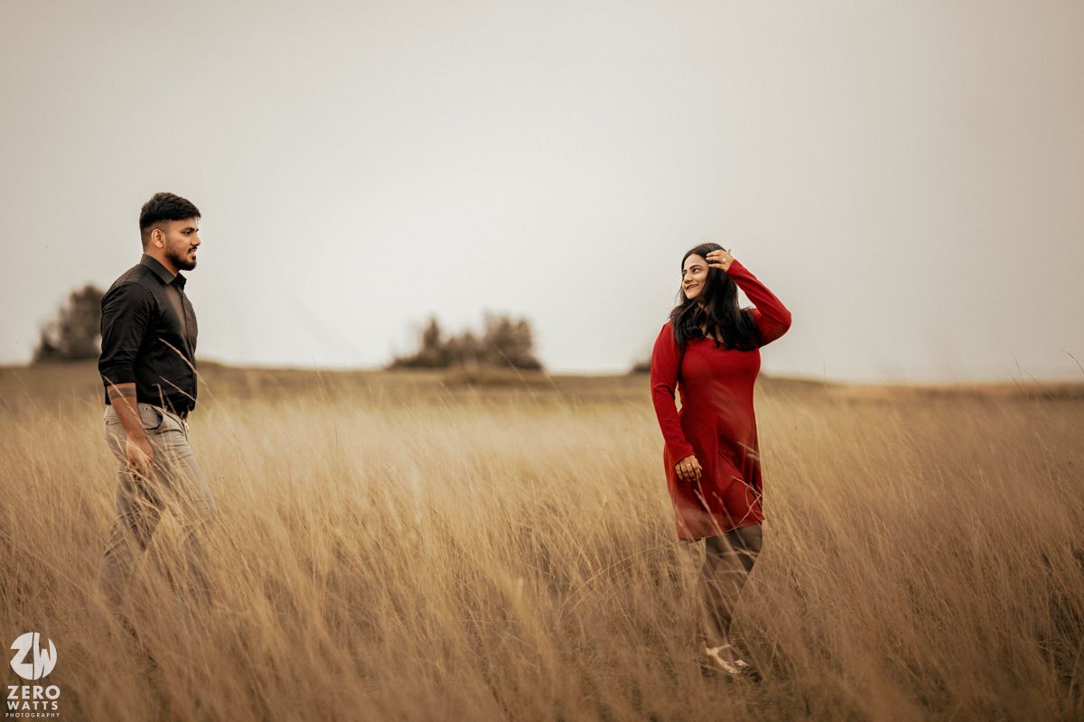 A couple poses playfully in a wheat field with the person in red looking through their hands.