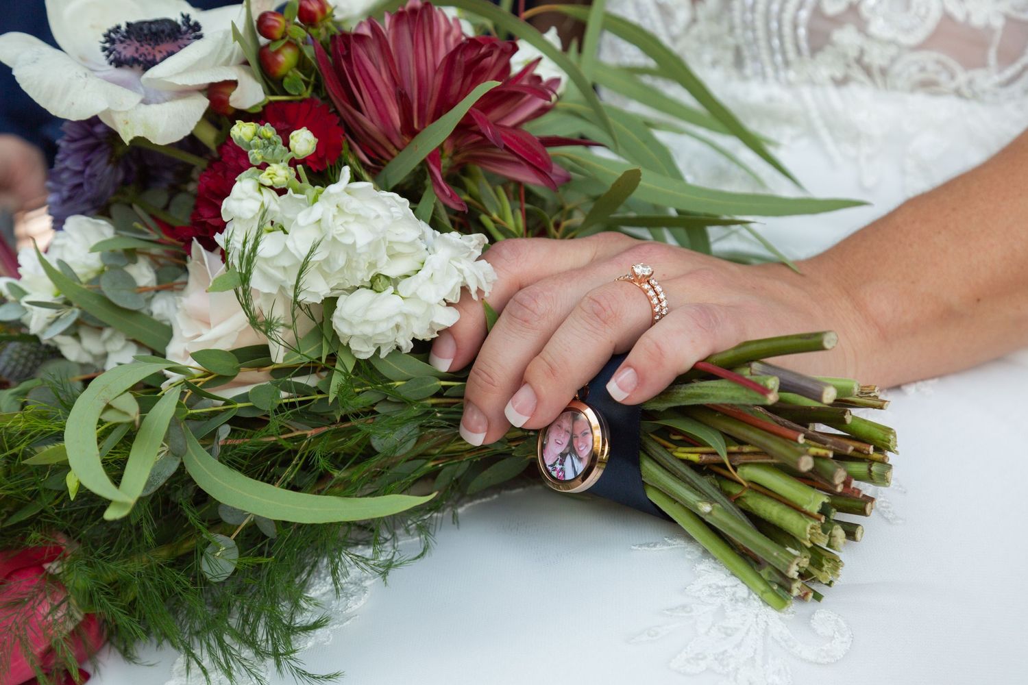 Hand with engagement ring holds elegant wedding bouquet with white and burgundy flowers wrapped in ribbon near lace tablecloth.