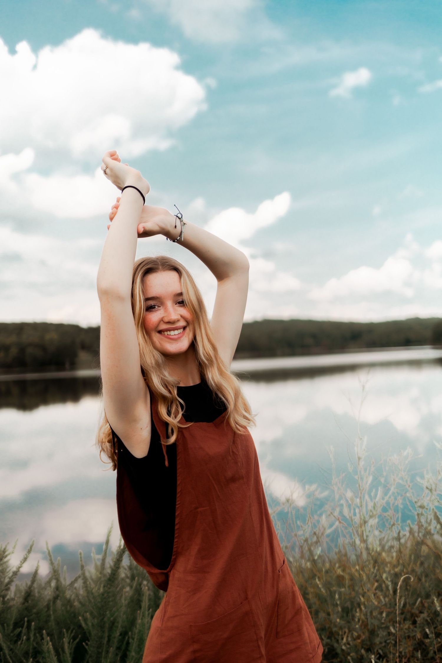 Joyful summer portrait by lake with arms raised against blue sky and clouds.