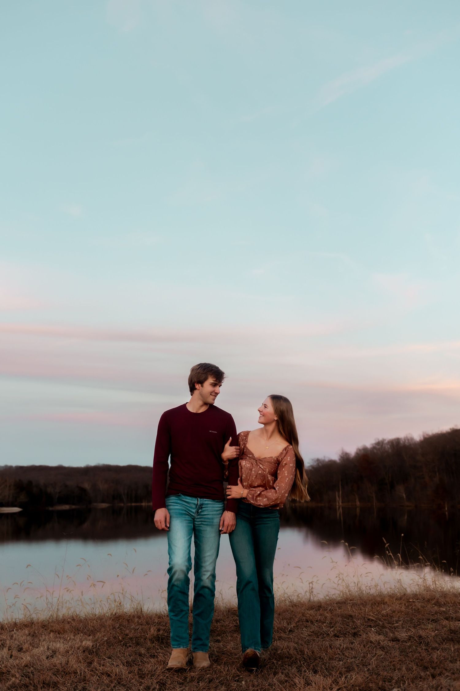 A couple stands together against a lakeside sunset backdrop.