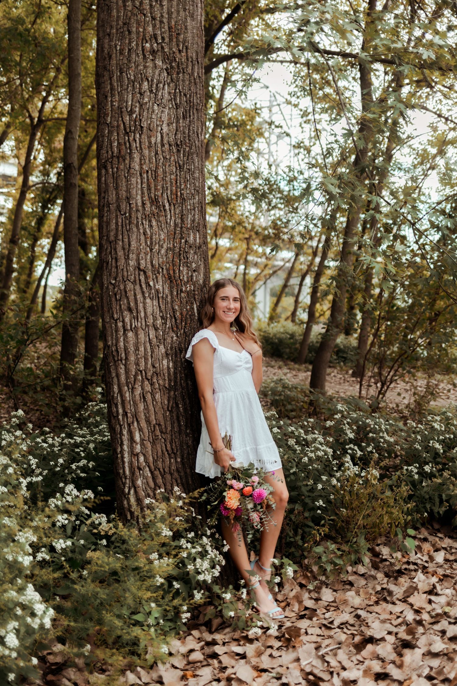 A figure in a white sundress poses against a large tree trunk surrounded by delicate white wildflowers in a forest setting.