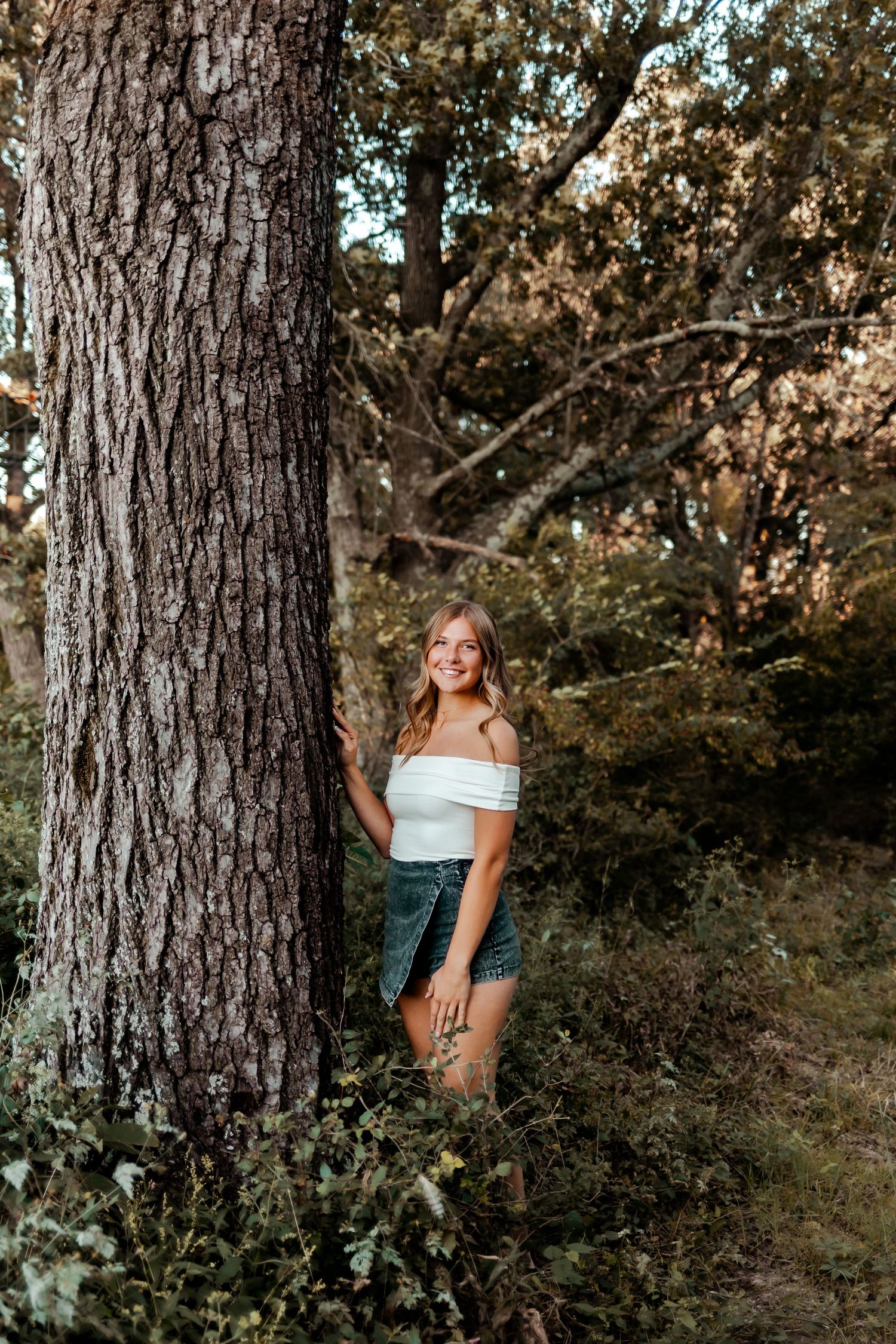Portrait of someone in a white off-shoulder top posing against a large tree trunk in a natural outdoor setting.