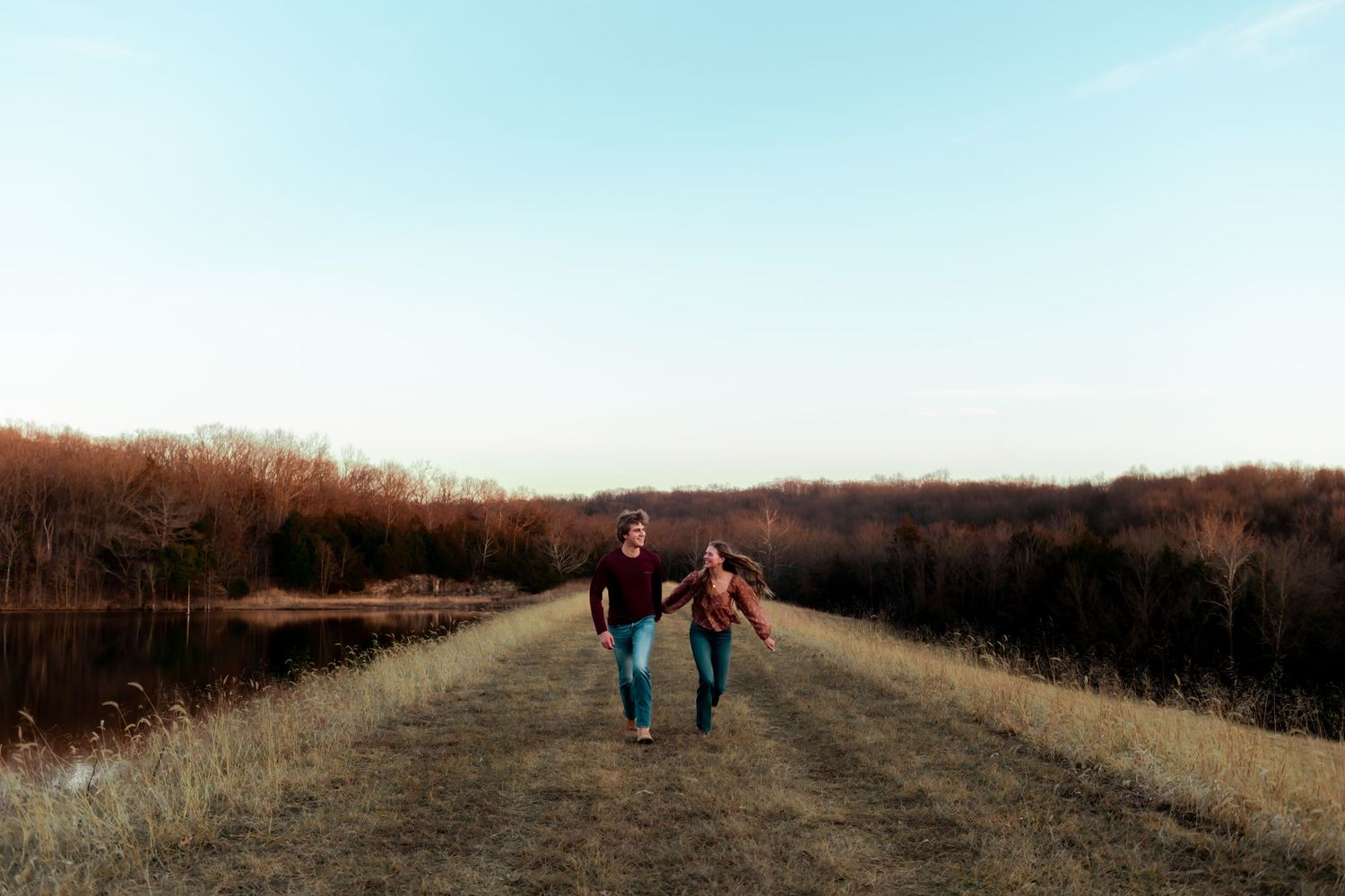 Two people walk hand in hand along a path near a serene lake at dusk.