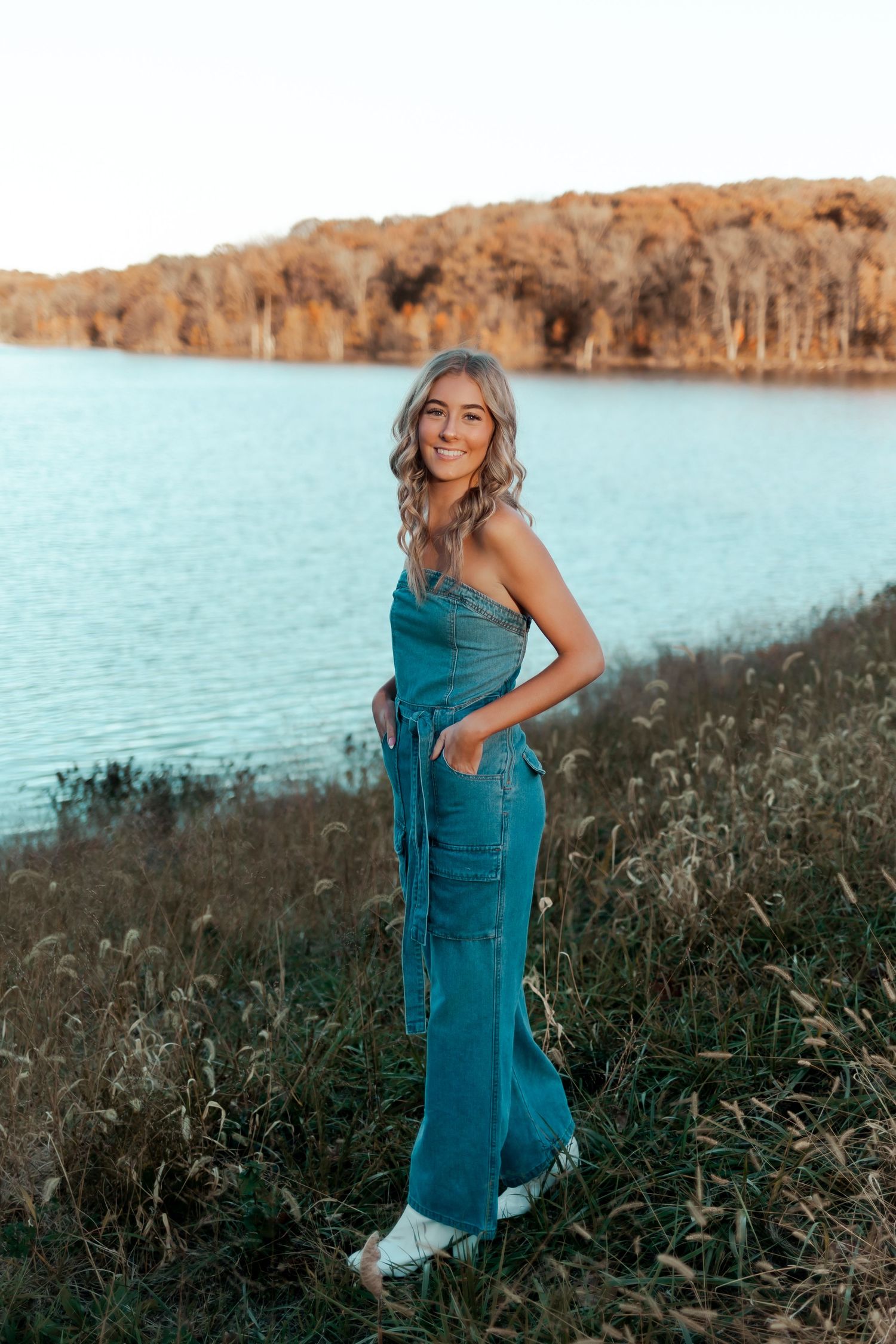 A person in teal jumpsuit poses on a hillside overlooking a turquoise lake.