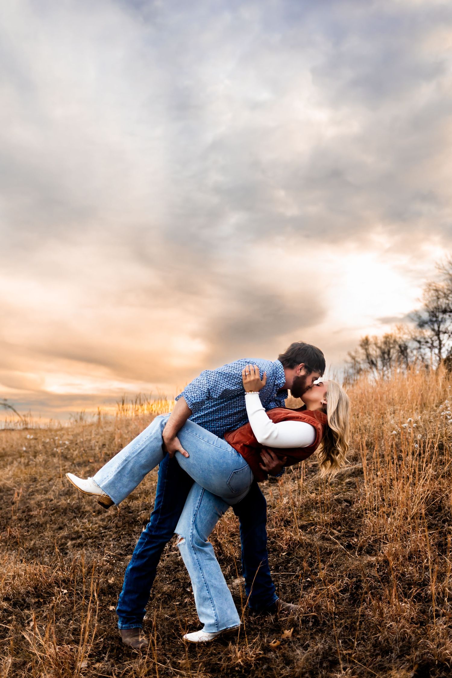 Romantic couple embraces in a golden field at sunset, creating a dramatic silhouette against the cloudy sky.