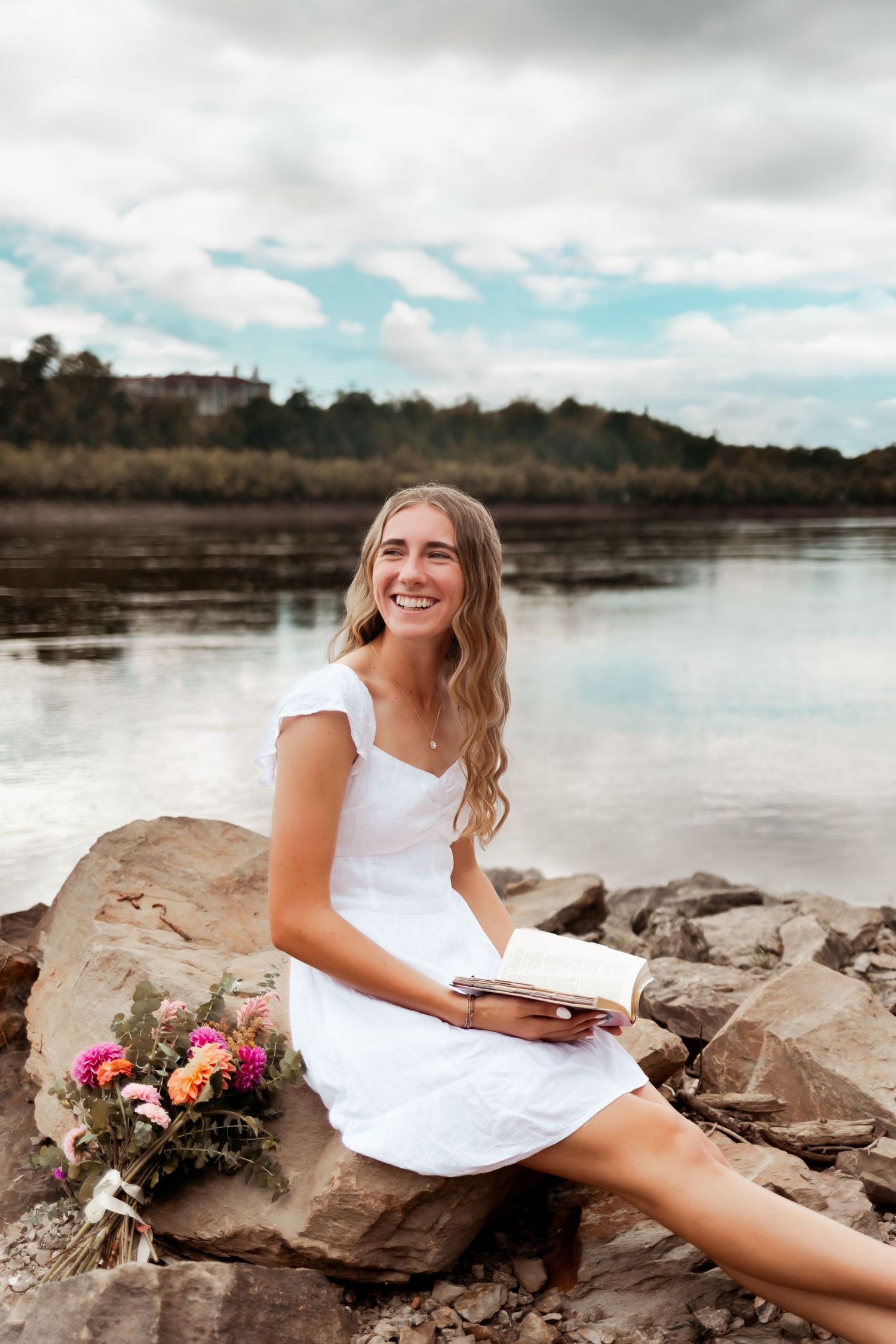 A figure in white dress sits on rocky shore holding book with colorful flower bouquet beside them.