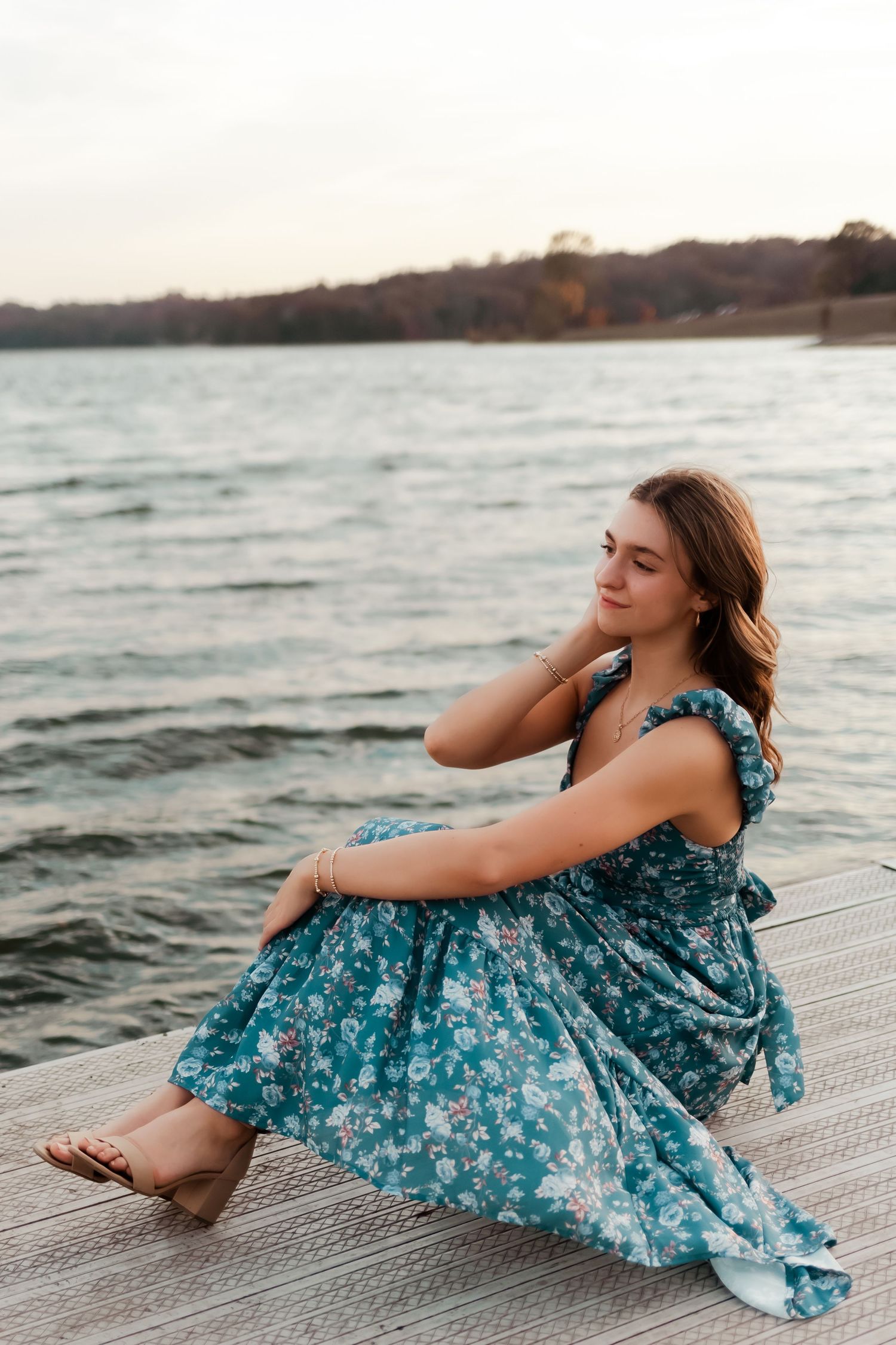 Person in floral blue dress sitting on dock by lake during sunset.