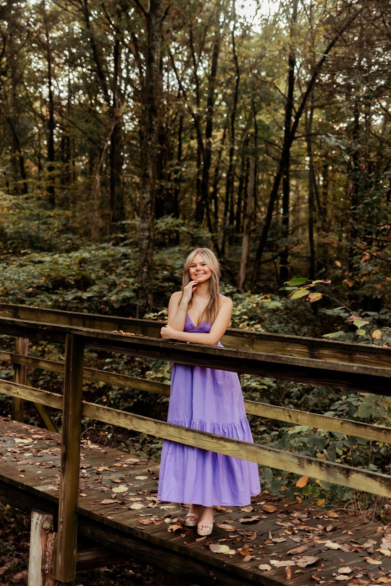 Person in purple maxi dress posing on wooden bridge in forest setting.