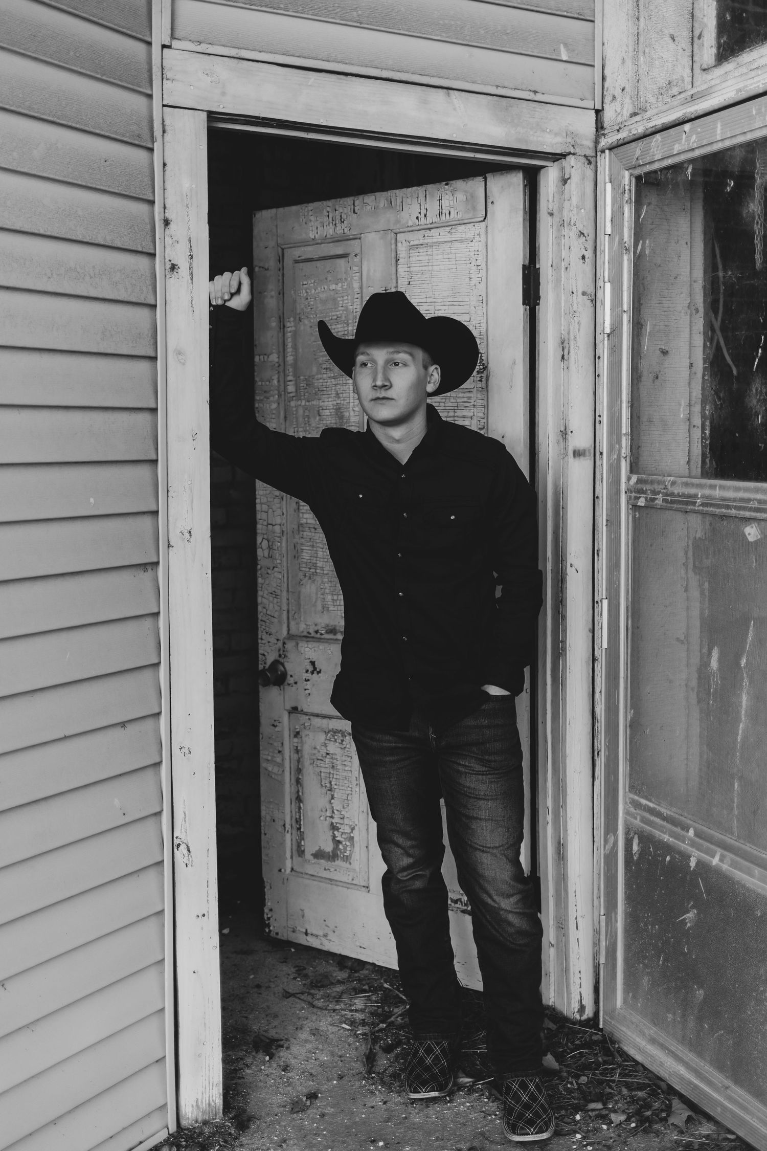 A black and white portrait of someone in cowboy hat standing in a rustic doorway.