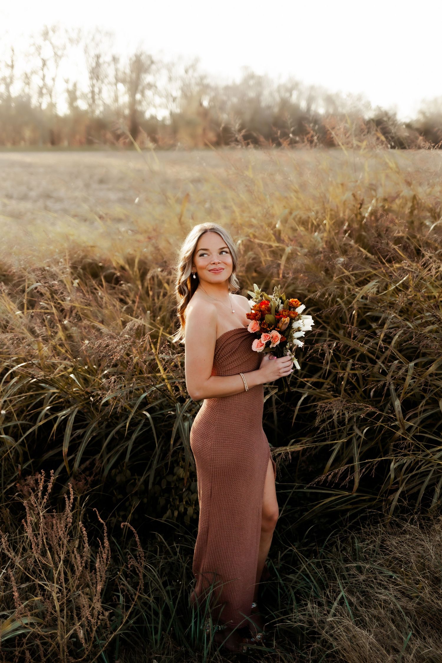 A figure in a brown dress holds a vibrant autumn bouquet while standing in a golden field at sunset.