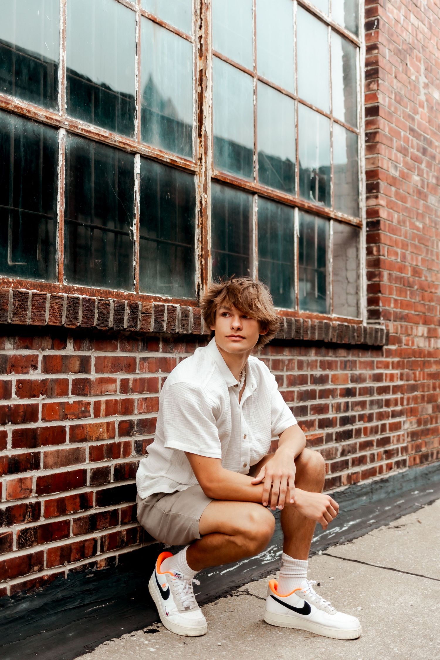 Someone in casual attire crouches by a brick wall with industrial windows.