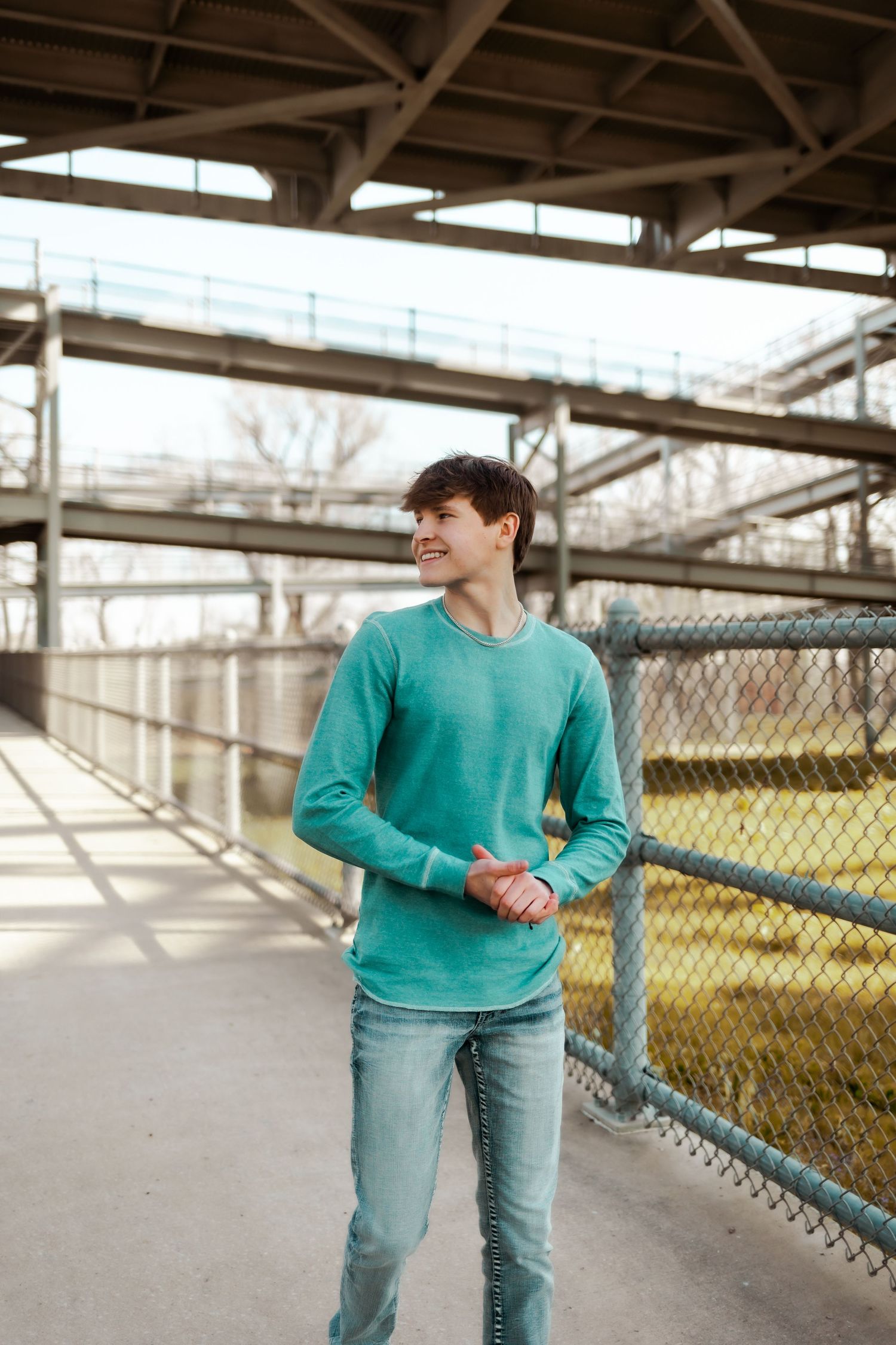 A person in a teal sweater and jeans smiles while standing near an industrial overpass structure.