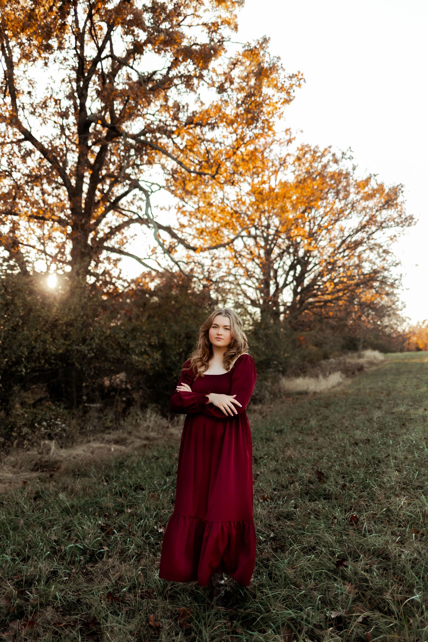 Person in burgundy maxi dress standing in autumn field at sunset with golden trees.