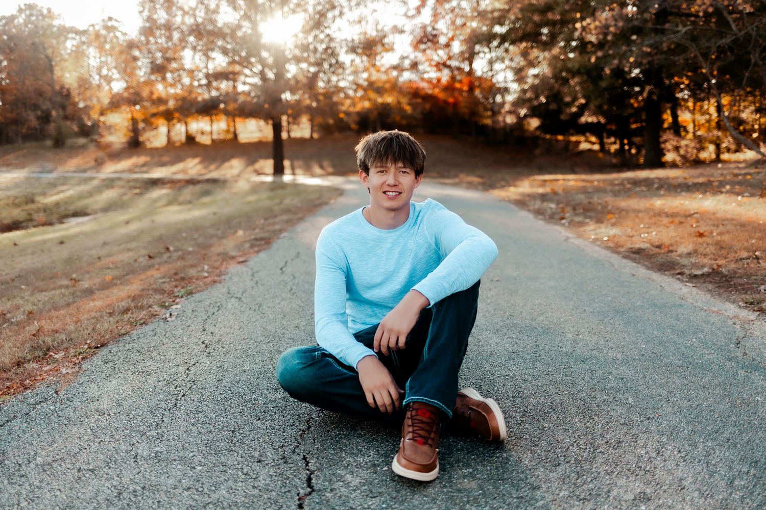 A person in blue sweater sits cross-legged on paved path during golden hour sunset.
