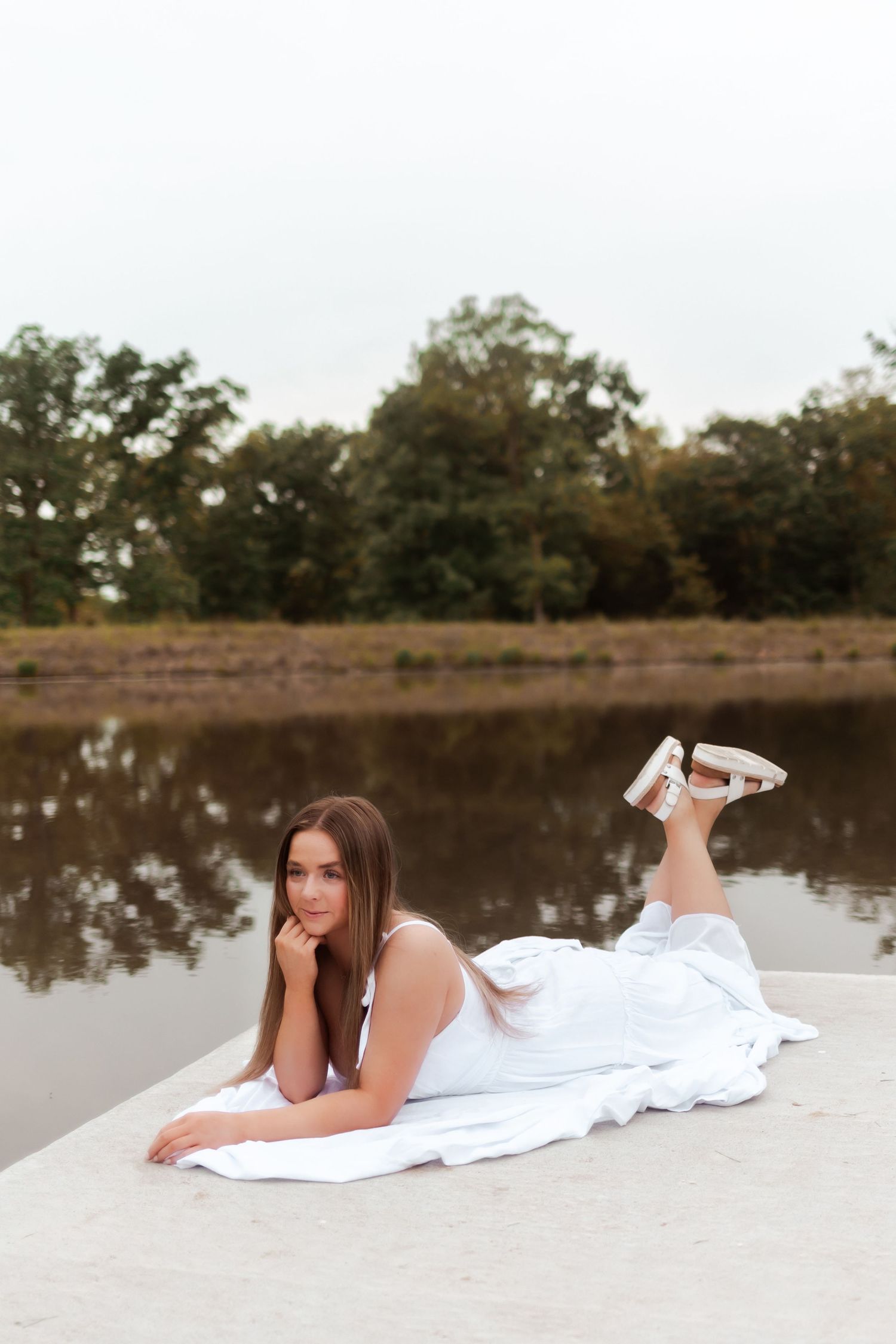 Elegant pose on concrete pier with reflective lake and trees in background.