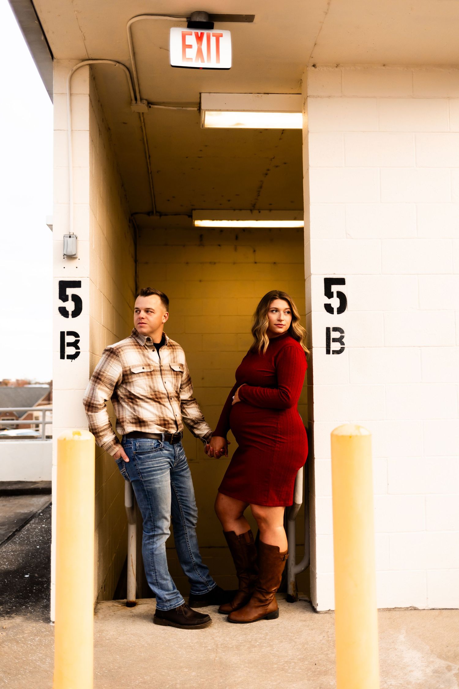 Couple poses in urban parking structure entrance with exit sign and numbered pillars.