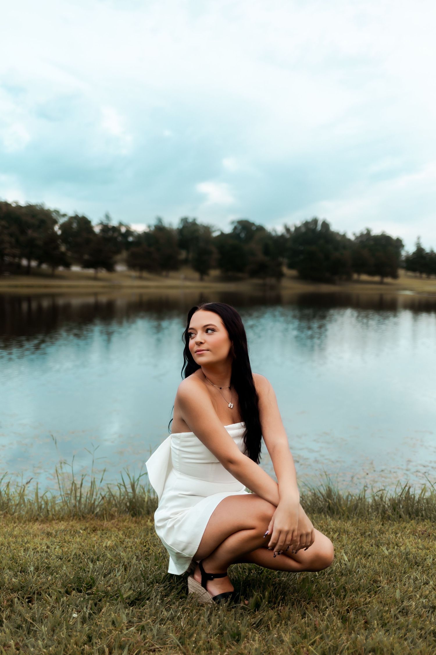 A figure in white dress crouches thoughtfully by calm lake under cloudy sky.