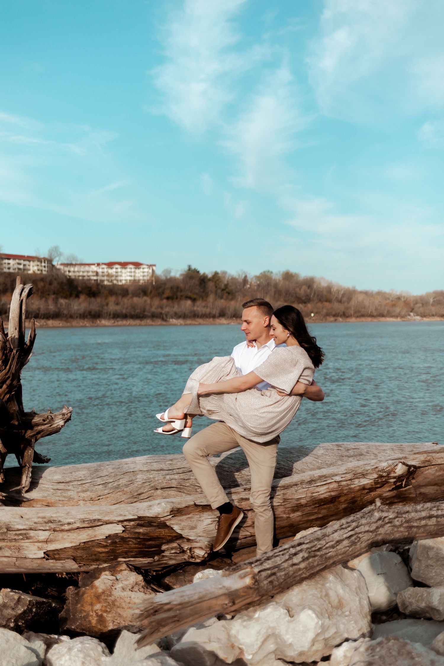 Couple shares playful moment while sitting on driftwood by riverside on sunny day.