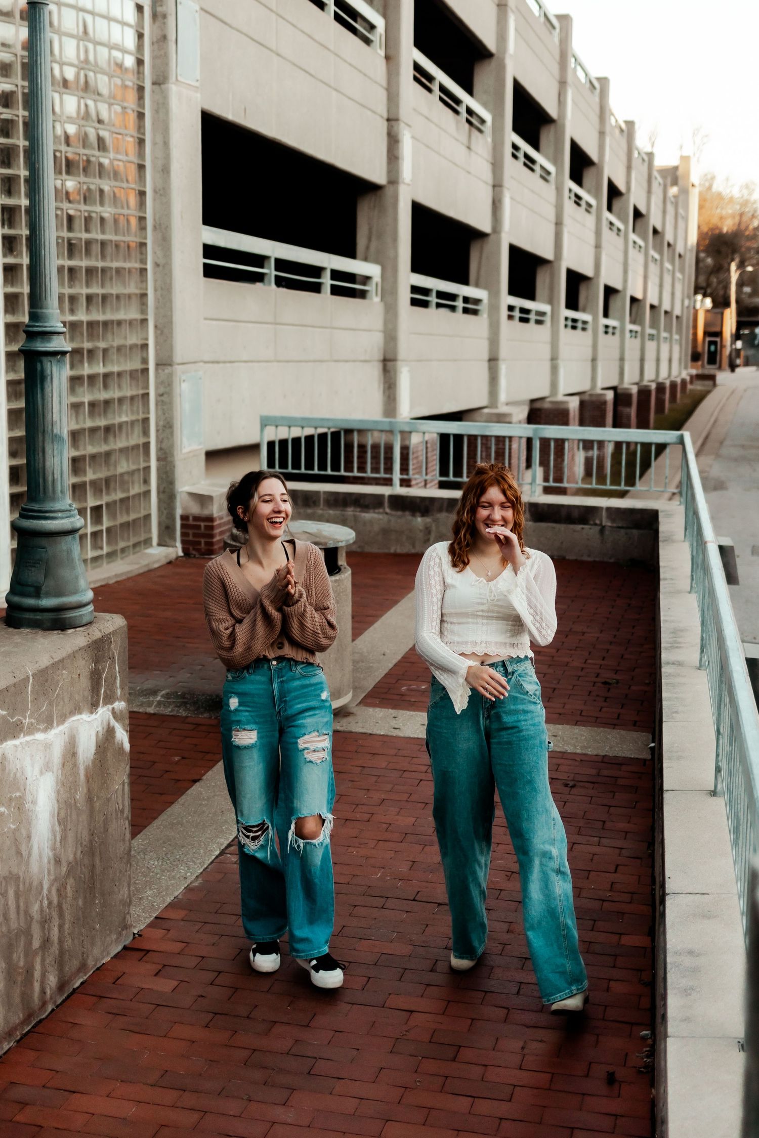 Two friends in retro-style jeans laugh while walking along brick pathway near parking structure.