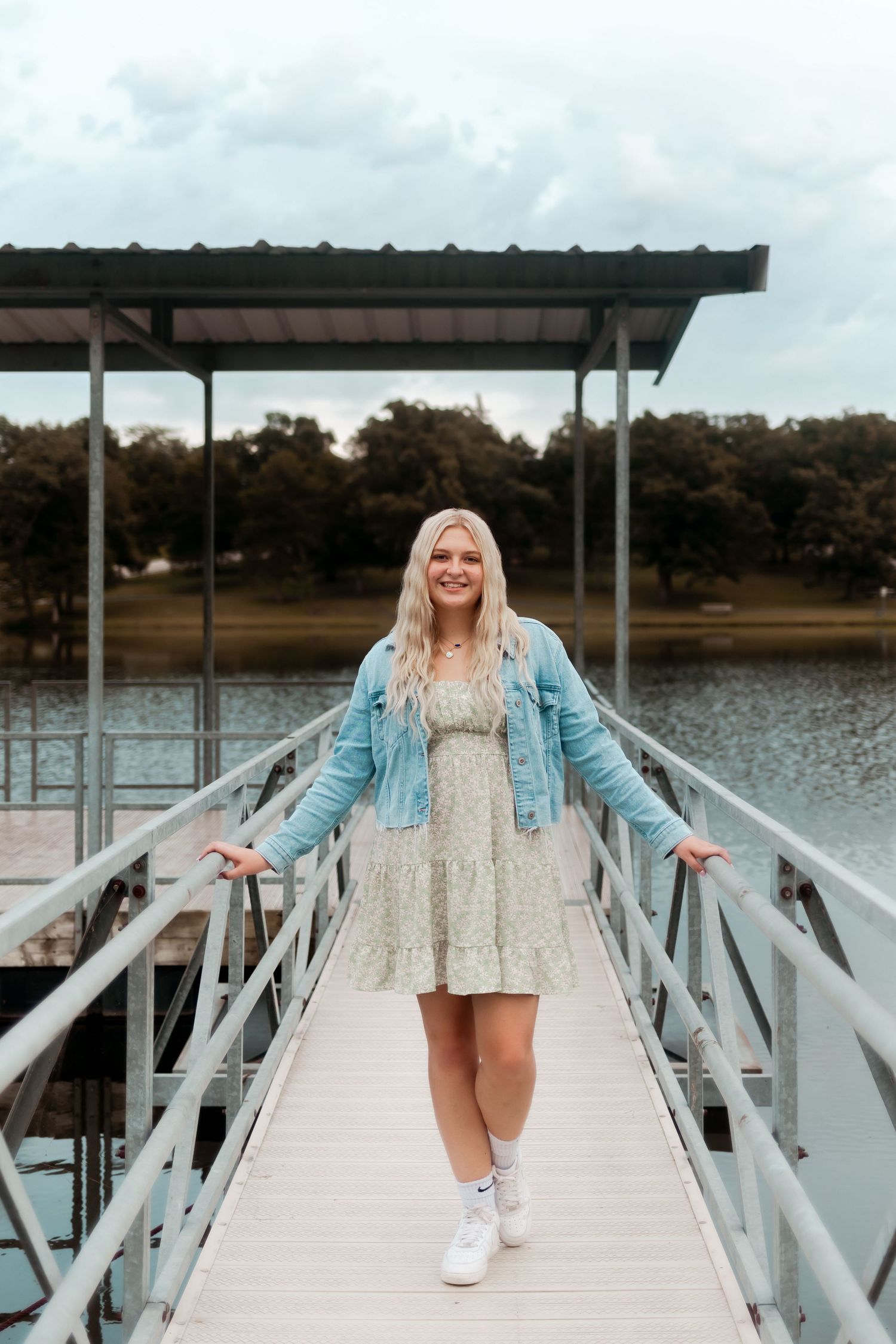 A figure in denim jacket and floral dress poses cheerfully on metal dock walkway over serene lake.