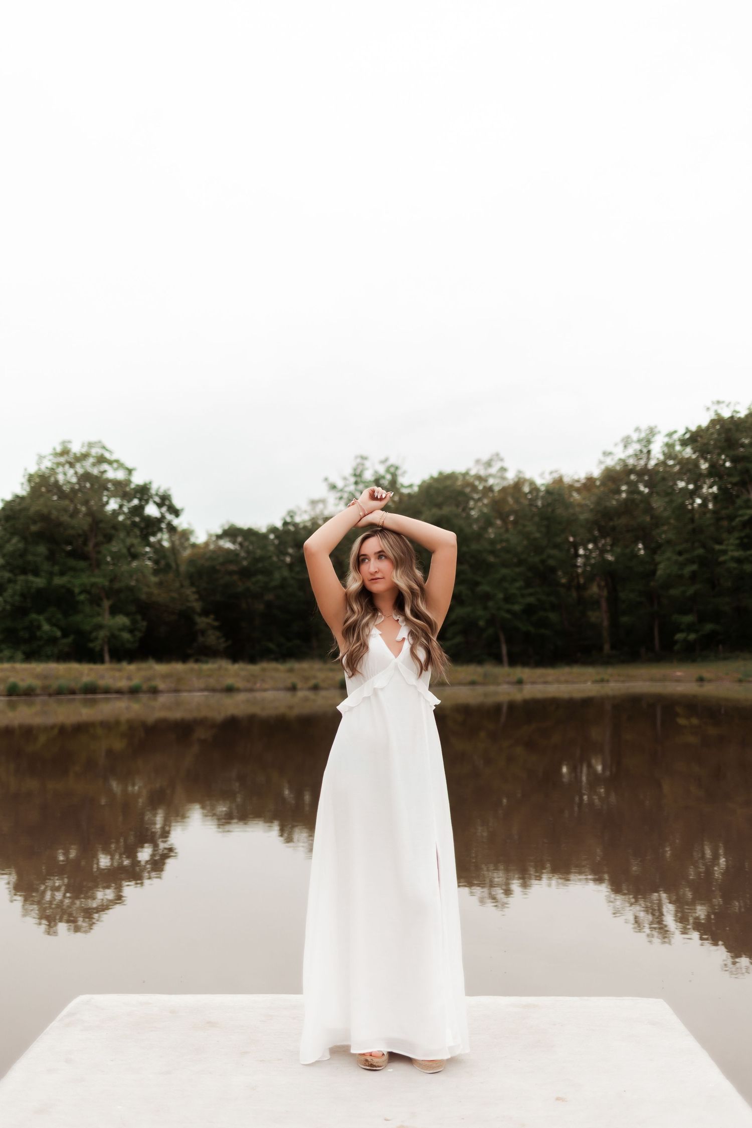 Fashion portrait of person in flowing white dress standing on dock by still lake.